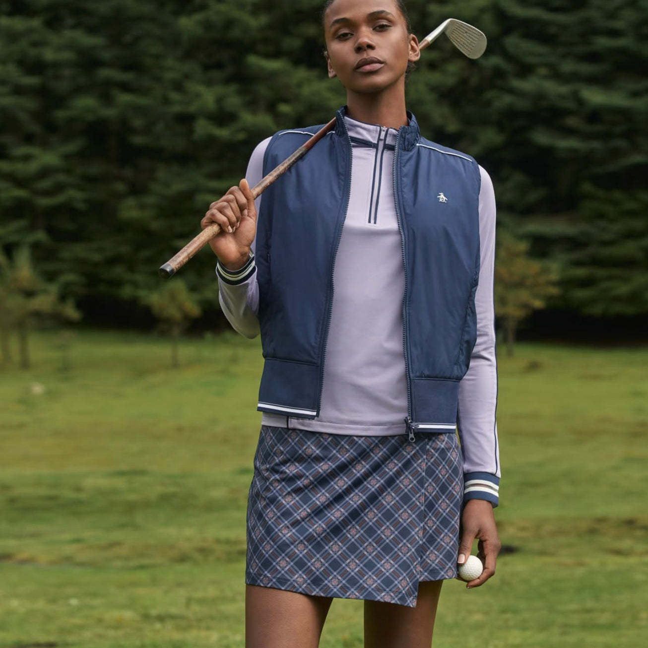 Female golfer wearing Original Penguin Ladies Plaid Wrap Golf Skort with Long Sleeve Sun Protection Top in lilac with her golf club resting over her shoulder.