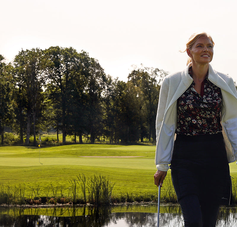 Woman walking on a golf course with trees and water in the background wearing Abacus golf wear including a light fleece jacket, floral print polo, navy skort and navy leggings.