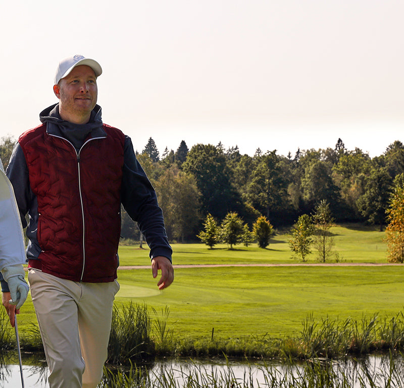 Man walking on a golf course with trees and water in the background wearing Abacus golf wear including a burgundy gilet over a blue hooded layer