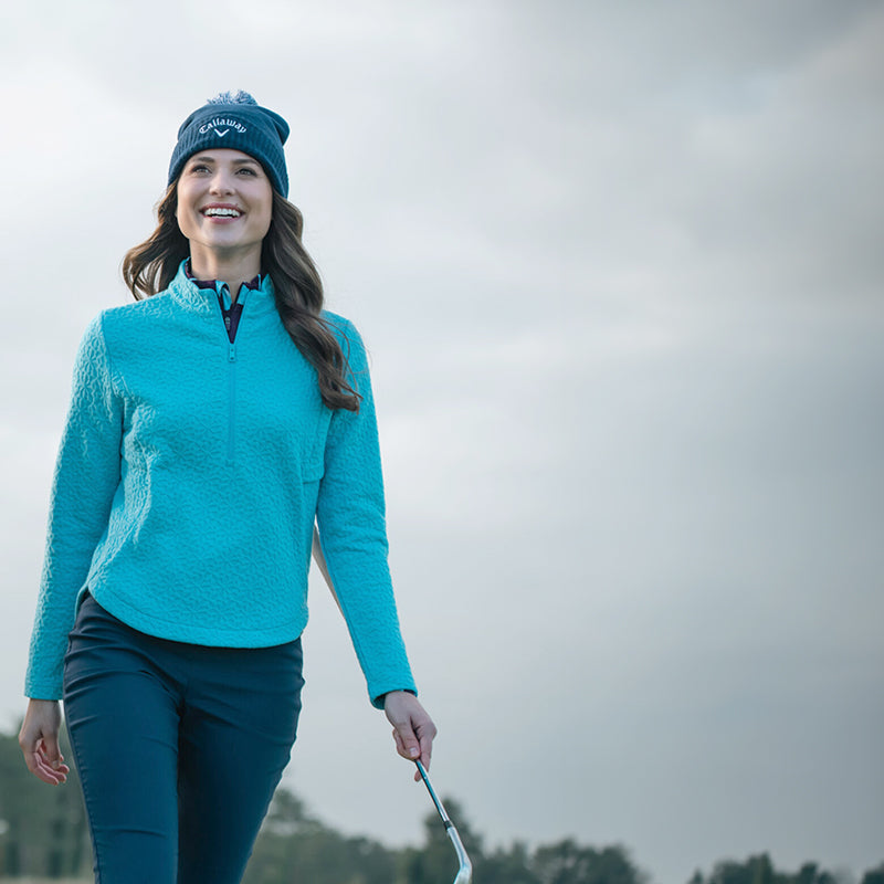 Female golfer holding a golf club and walking wearing a navy Callaway bobble hat and bright, ethereal blue textured zip neck mid layer