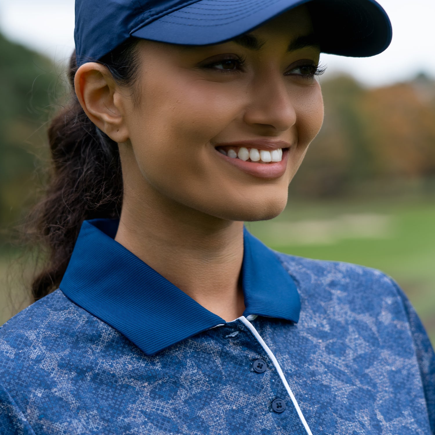 A close-up of a smiling woman wearing a blue floral print textured polo shirt and a matching blue cap, from the Callaway golf ladies high summer and autumn winter collection.