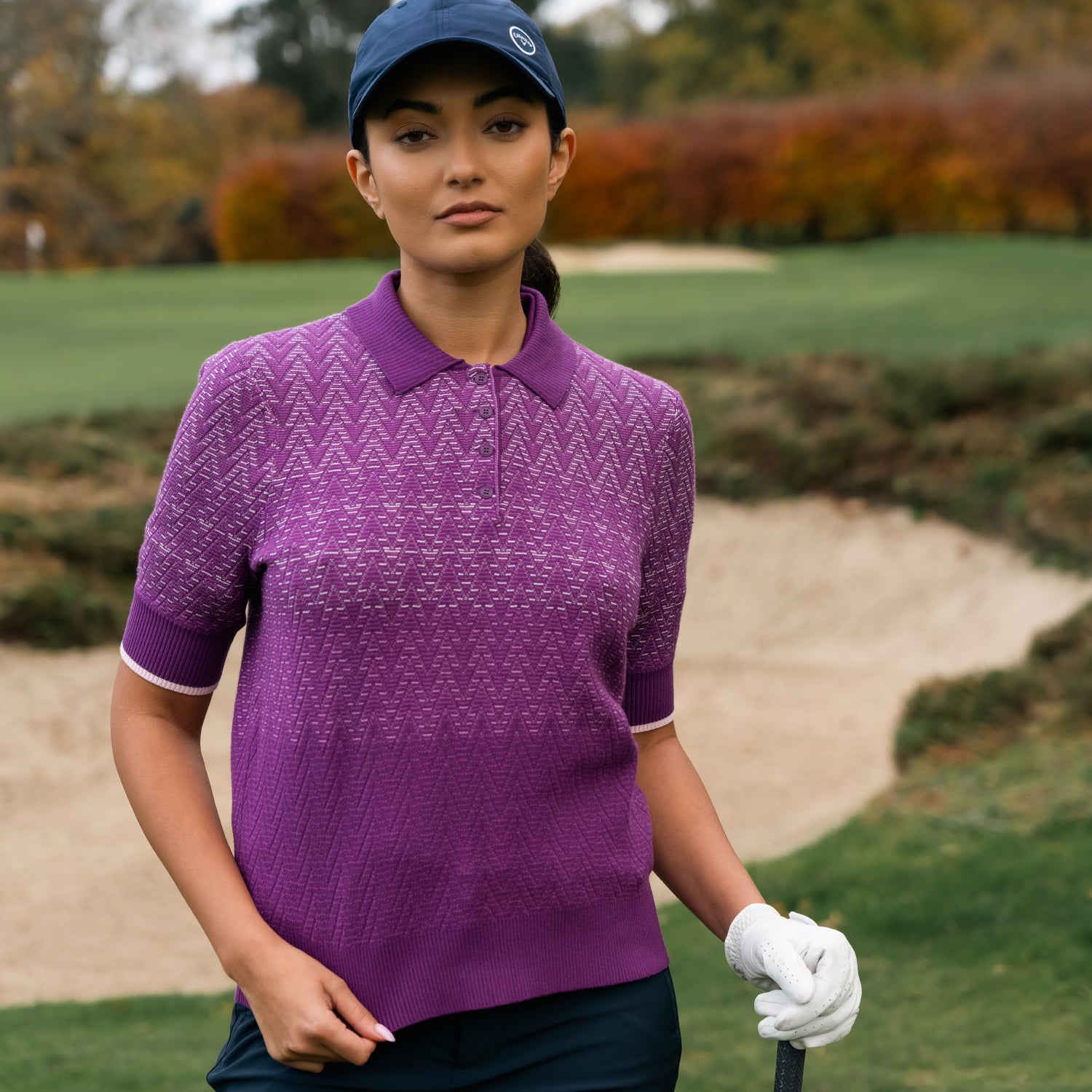 A woman on a golf course wearing a rich plum short-sleeve knit polo shirt with a graduated geometric pattern from Callaway, a navy blue golf cap, a white golf glove, and navy trousers.