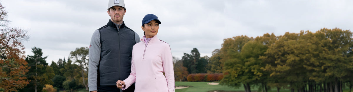 A man and a woman standing on a golf course wearing Callaway Golf autumn winter 2026 clothing. The man wears a white cap, a dark gray zip-up gilet over a light gray long-sleeve shirt, and black pants. The female golfer wears a navy Callaway cap and light pink mid layer quarter zip with contrast piping.