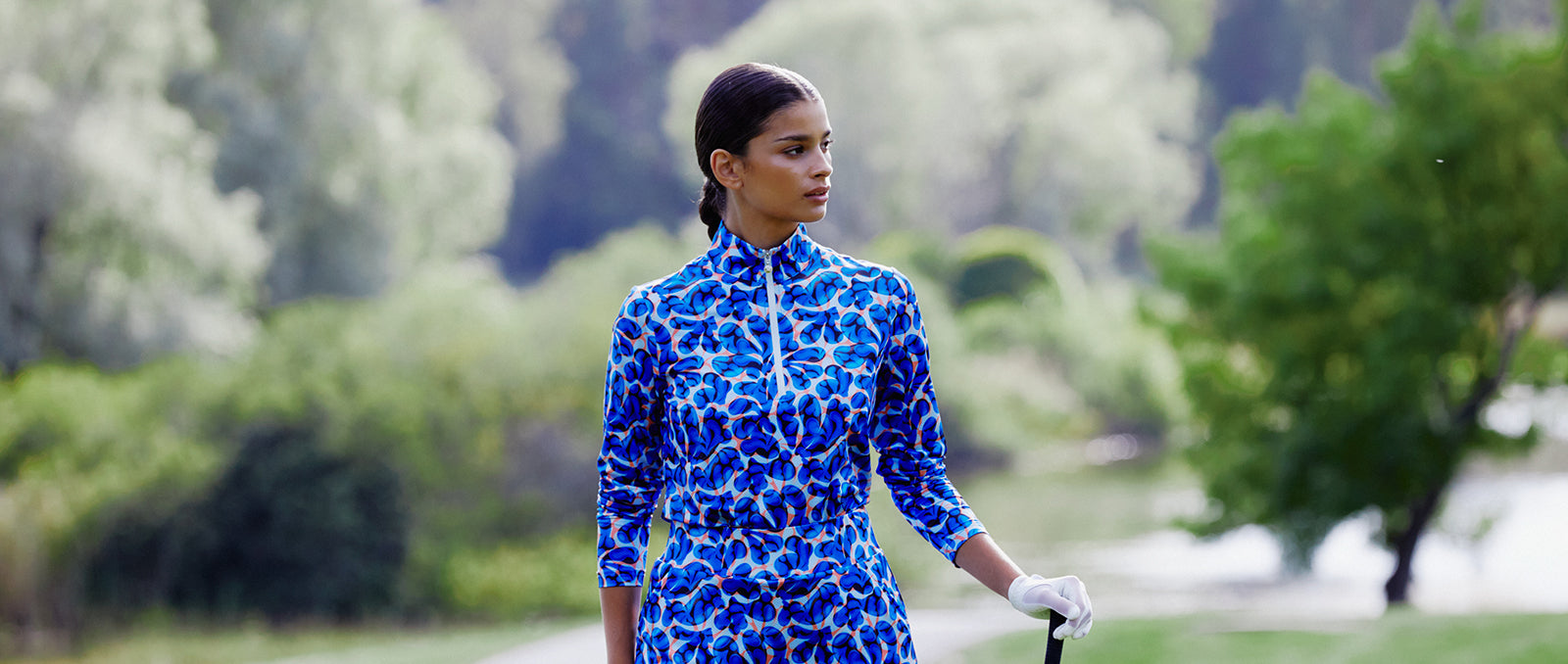 Woman golfer holding a club wearing Daily Sports abstract floral print zip neck top and skort from the spring summer 2026 collection at GolfGarb
