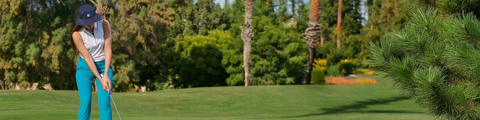 Woman playing golf completing her shot wearing the FootJoy navy bucket sun hat , sleeveless polo and vibrant blue 7-8th cropped rousers with trees in the background