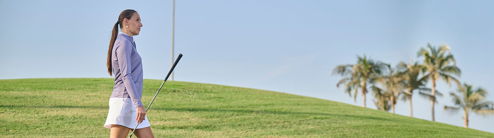 Female golfer walking across a fairway wearing a FootJoy golf zip neck top in soft lilac and a white pull on skort