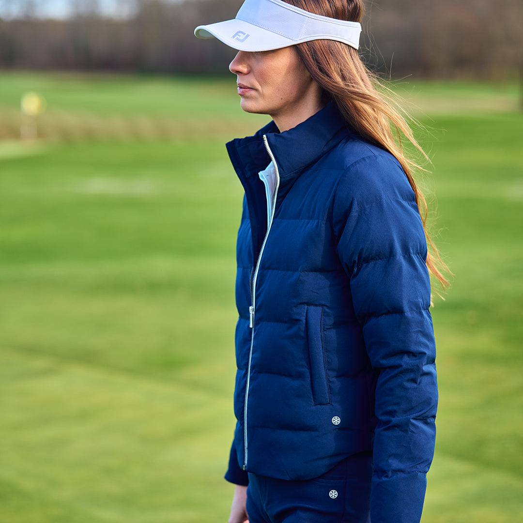Woman in a FootJoy blue padded insulated jacket and white visor standing on a golf course.