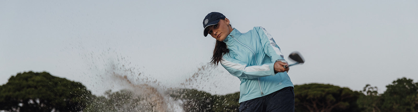 Female golfer wearing the pastel blue Galvin Green Pertex waterproof golf jacket taking a shot from a bunker