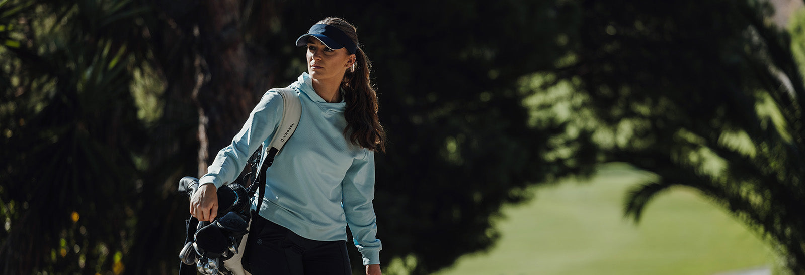 Female golfer walking to the next hole with her club bag over her shoulder and wearing the Galvin Green pale blue hooded sweatshirt and navy visor