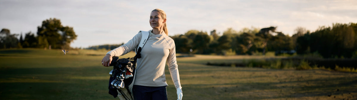 Female golfer on a golf course, club bag over her shoulder wearing the Galvin Green ladies autumn winter 2026 2027 Insula thermal hoodie in sand
