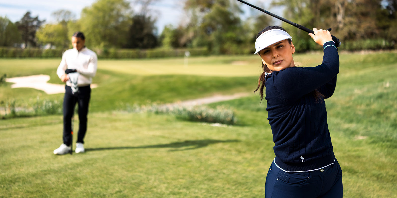 Female golfer swinging her club wearing Glenmuir women's navy v neck with white trim sweater