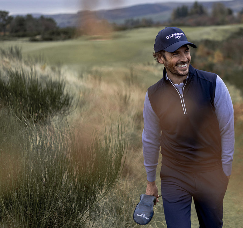 Man walking on a golf course wearing a cap and holding a golf club wearing Glenmuir men's navy golf trousers, navy gilet and powder blue sweater.