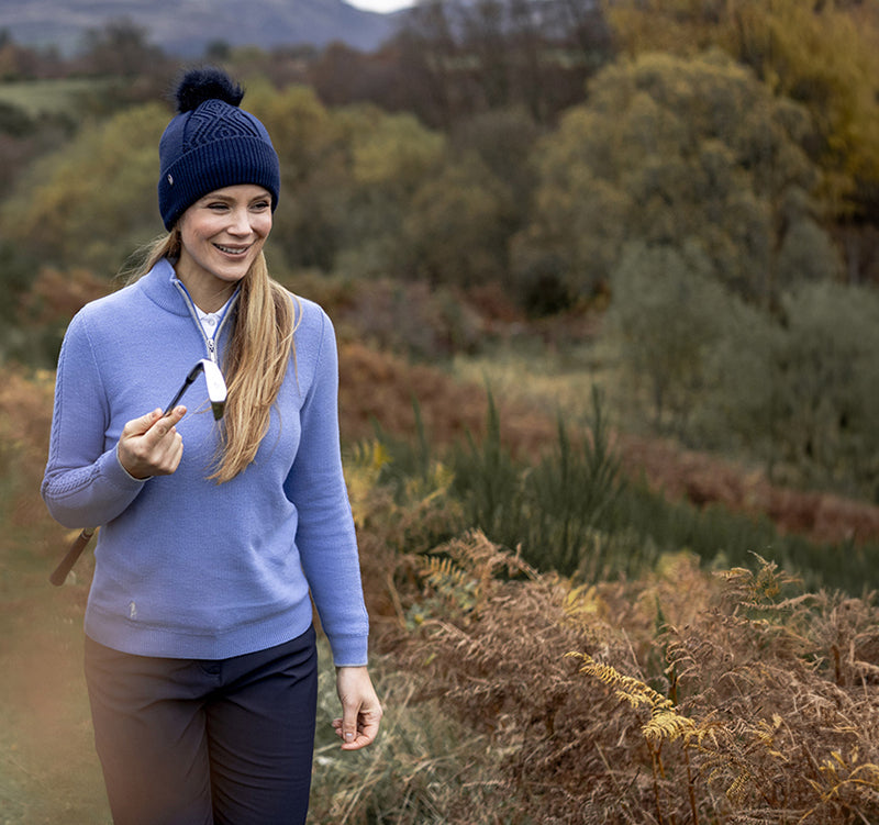 Woman golfer in a Glenmuir powder blue sweater, navy golf trousers and navy knit bobble hat holding a golf club as she walks to the next tee.