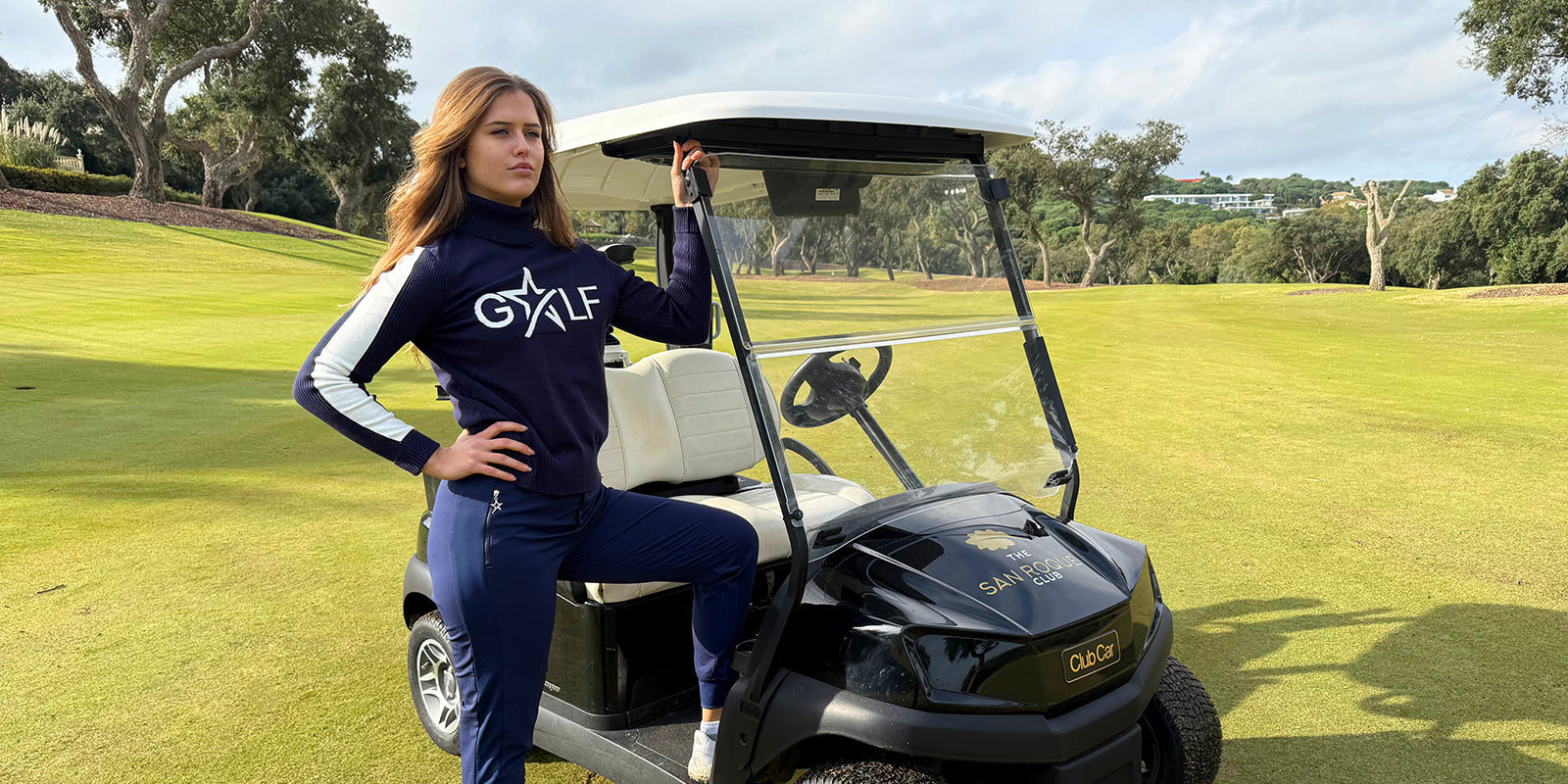 Woman in Swing Out Sister golf jumper and jogger style trousers in blue, standing next to a golf cart on a golf course