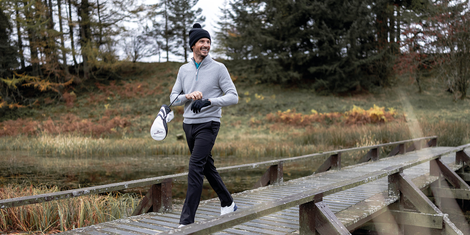 Man walking across a wooden bridge at a golf course wearing black Glenmuir trousers, grey Glenmuir sweater, black Glenmuir bobble hat.