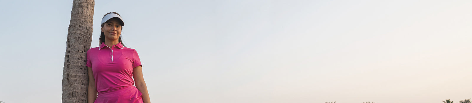 Female golfer leaning on the trunk of a palm tree wearing a vibrant pink Puma Golf ladies short sleeve polo with ribbed collar and white Puma Golf ladies visor
