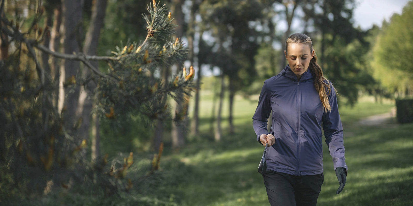 Female golfer walking past pine trees to the next tee on a golf course wearing Sunderland of Scotland women's Whisperdry waterproof jacket with hood.