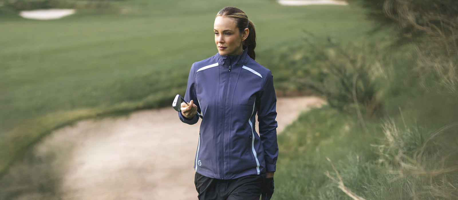 Female golfer walking past a bunker holding a golf club and wearing Sunderland of Scotland's Midnight Plum Killy waterproof jacket